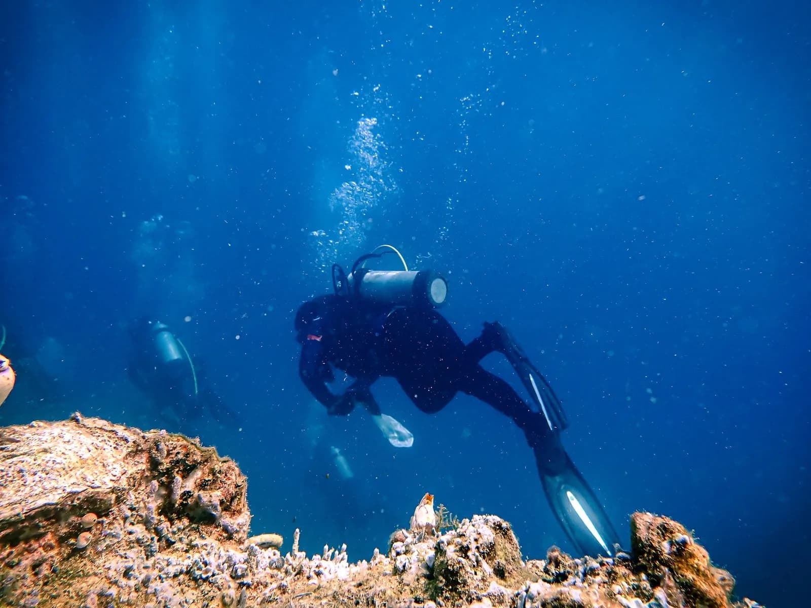 Plongeur sous-marin en combinaison explorant un récif corallien dans une eau tropicale bleue, un second plongeur en arrière-plan