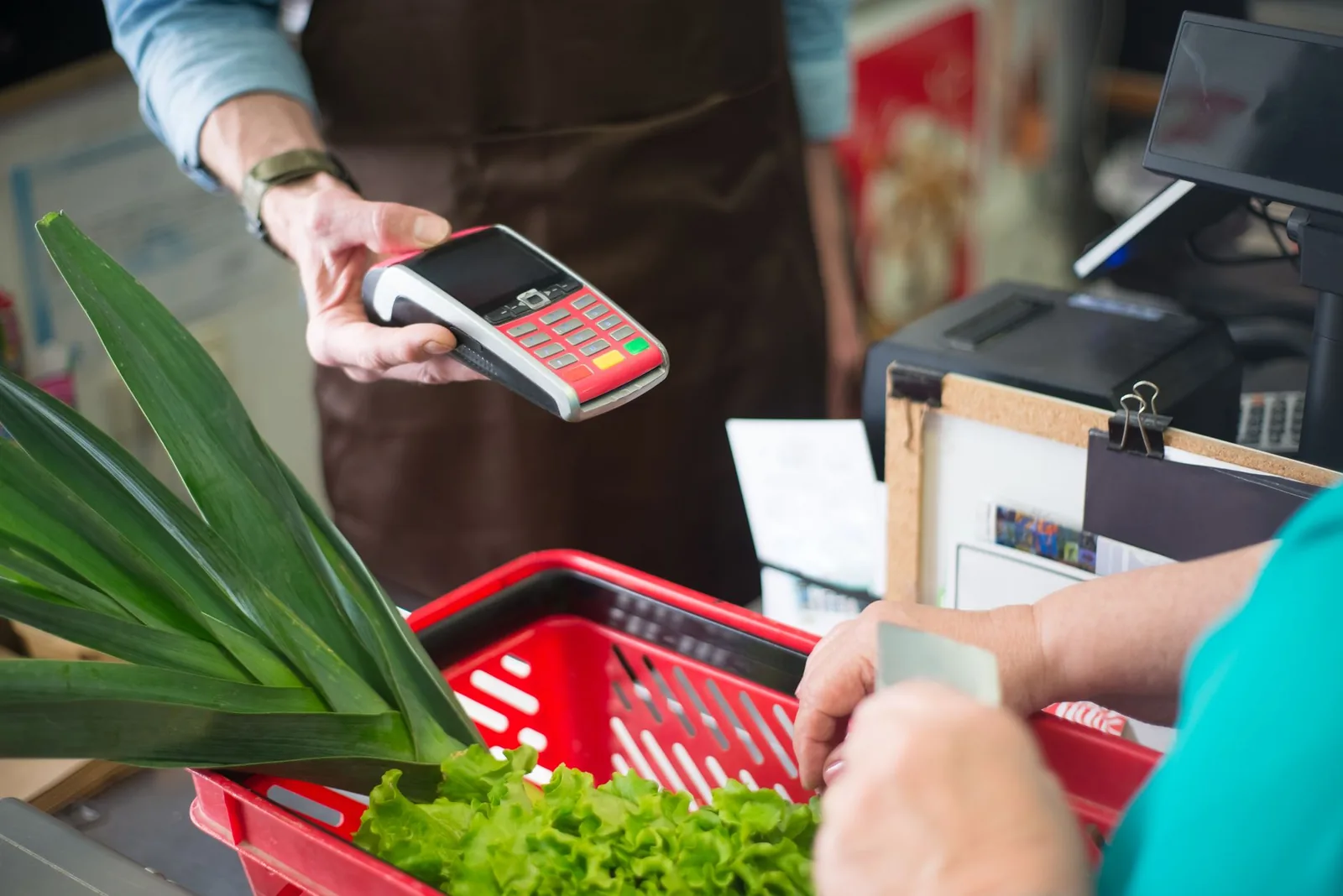 Paiement par carte à la caisse d'un supermarché, avec légumes frais (poireau, salade) dans le panier, illustrant le poste alimentation du budget mensuel à Nouméa