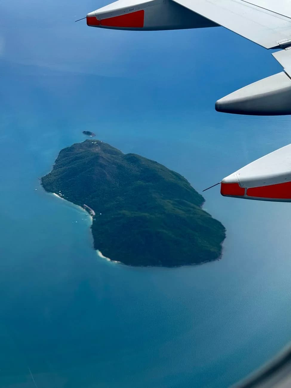Aile d'avion survolant une île tropicale verdoyante au milieu d'un océan bleu, vue depuis un hublot