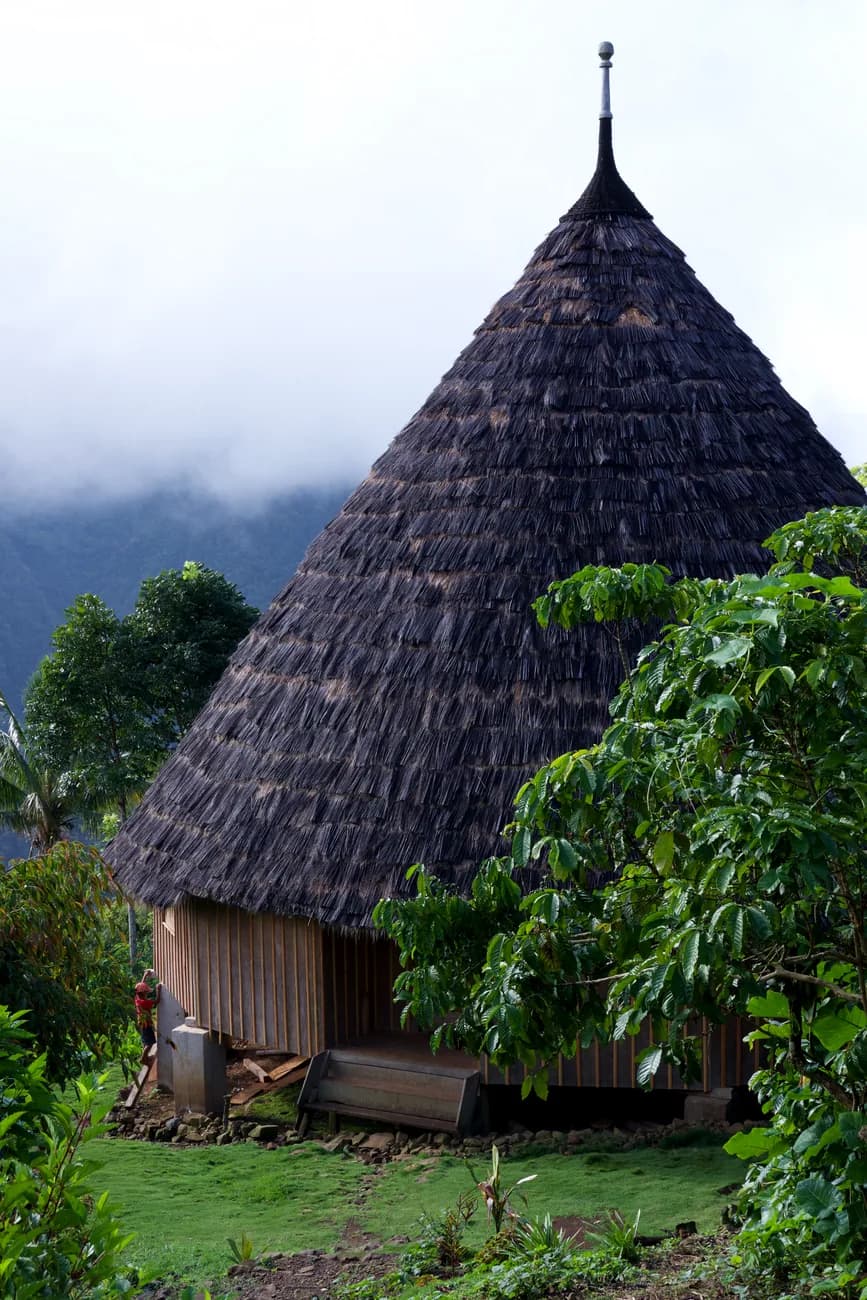 Case traditionnelle kanak au toit conique de chaume, sur pilotis de bois, dans un décor de montagnes et de végétation tropicale