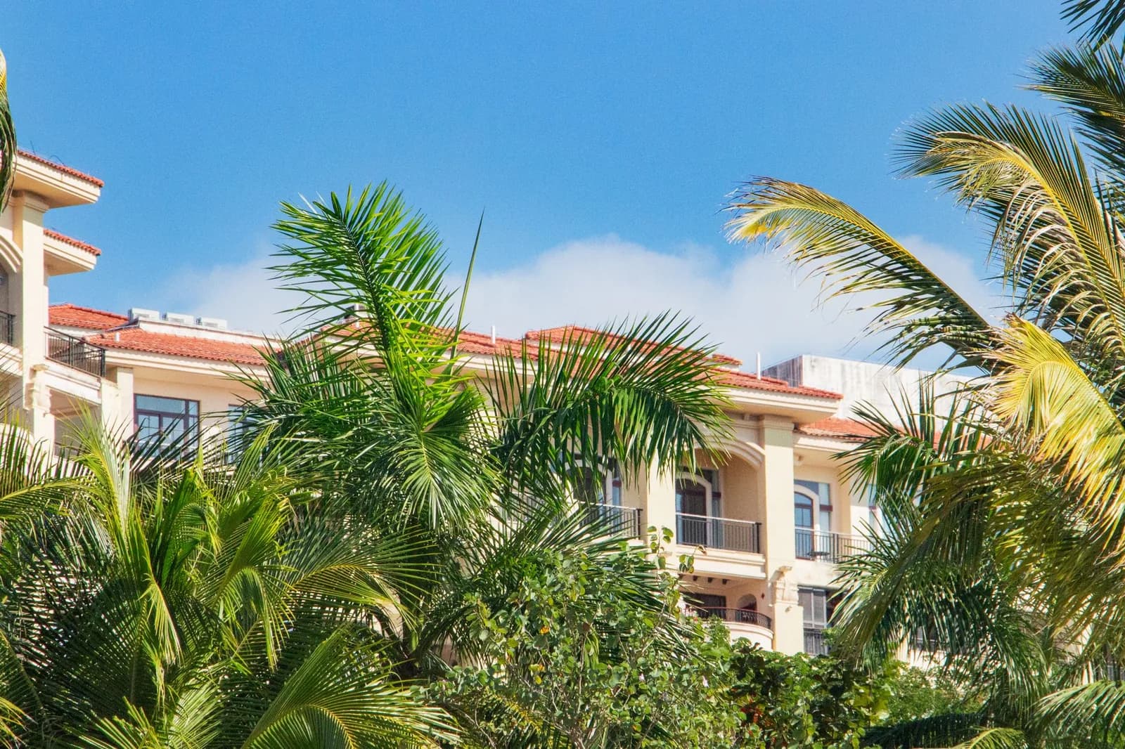 Immeuble résidentiel aux balcons et toiture de tuiles rouges, entouré de palmiers sous un ciel tropical
