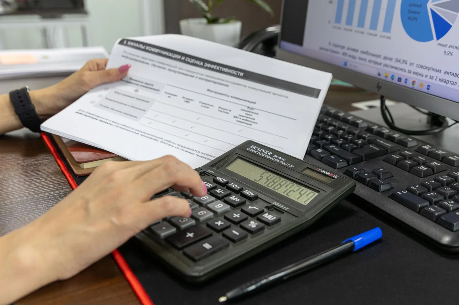 Mains manipulant des documents et une calculatrice sur un bureau, devant un ordinateur (image générique d'illustration)